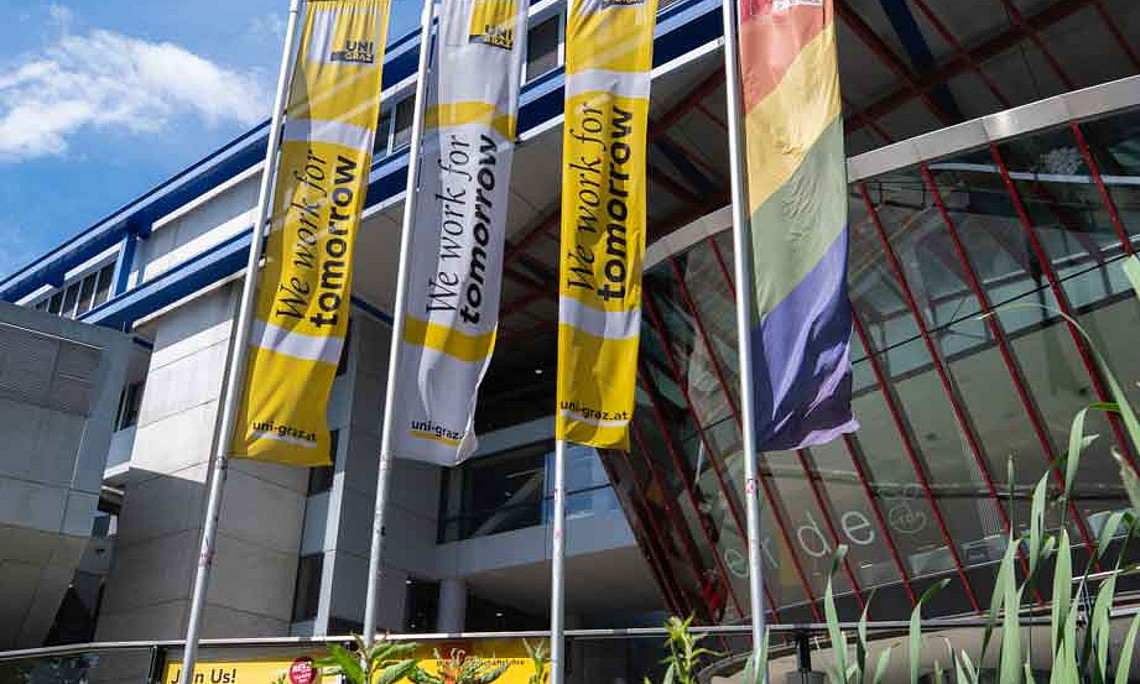 Drei Fahnen vor einem modernen Gebäude, zwei mit gelbem Schriftzug „We work for tomorrow“, eine in Regenbogenfarben.