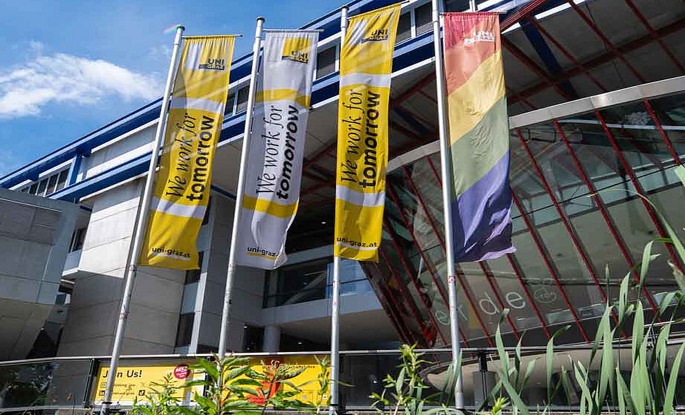 Drei Fahnen vor einem modernen Gebäude, zwei mit gelbem Schriftzug „We work for tomorrow“, eine in Regenbogenfarben.