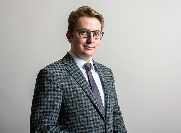 Junger Mann mit Brille, kariertem Sakko, weißem Hemd und Krawatte steht vor neutralem Hintergrund und blickt in die Kamera. ©P. Kalcher