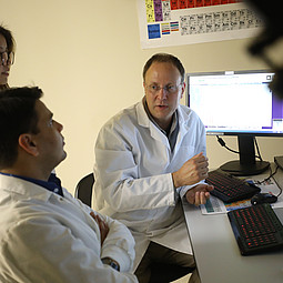 Fotografie von Bernd Zechmann bei der Arbeit an einem Schreibtisch mit Computern. Er trägt einen Laborkittel und erklärt gerade zwei anderen Personen, ebenfalls im Laborkittel, etwas.