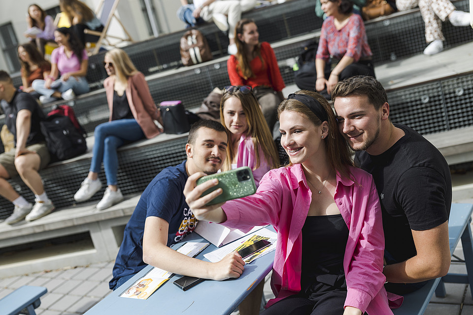 Studierende sitzen im Freien, eine Gruppe macht ein Selfie.