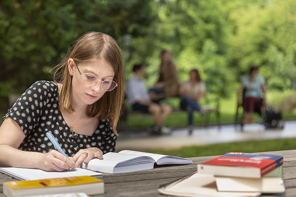 Eine Studierende sitzt im Freien und schreibt sich Notizen zu einem Buch auf.