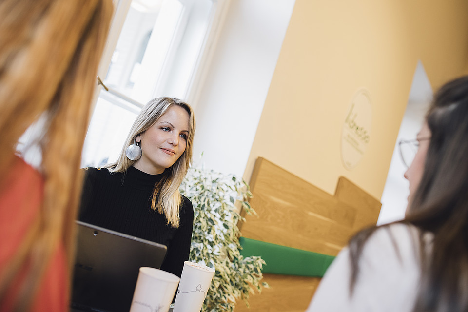 3 Studierende sitzen in einem Café; der Fokus ist auf die Studierende gerichtet, die in der MItte sitzt und einen Laptop vor sich stehen hat