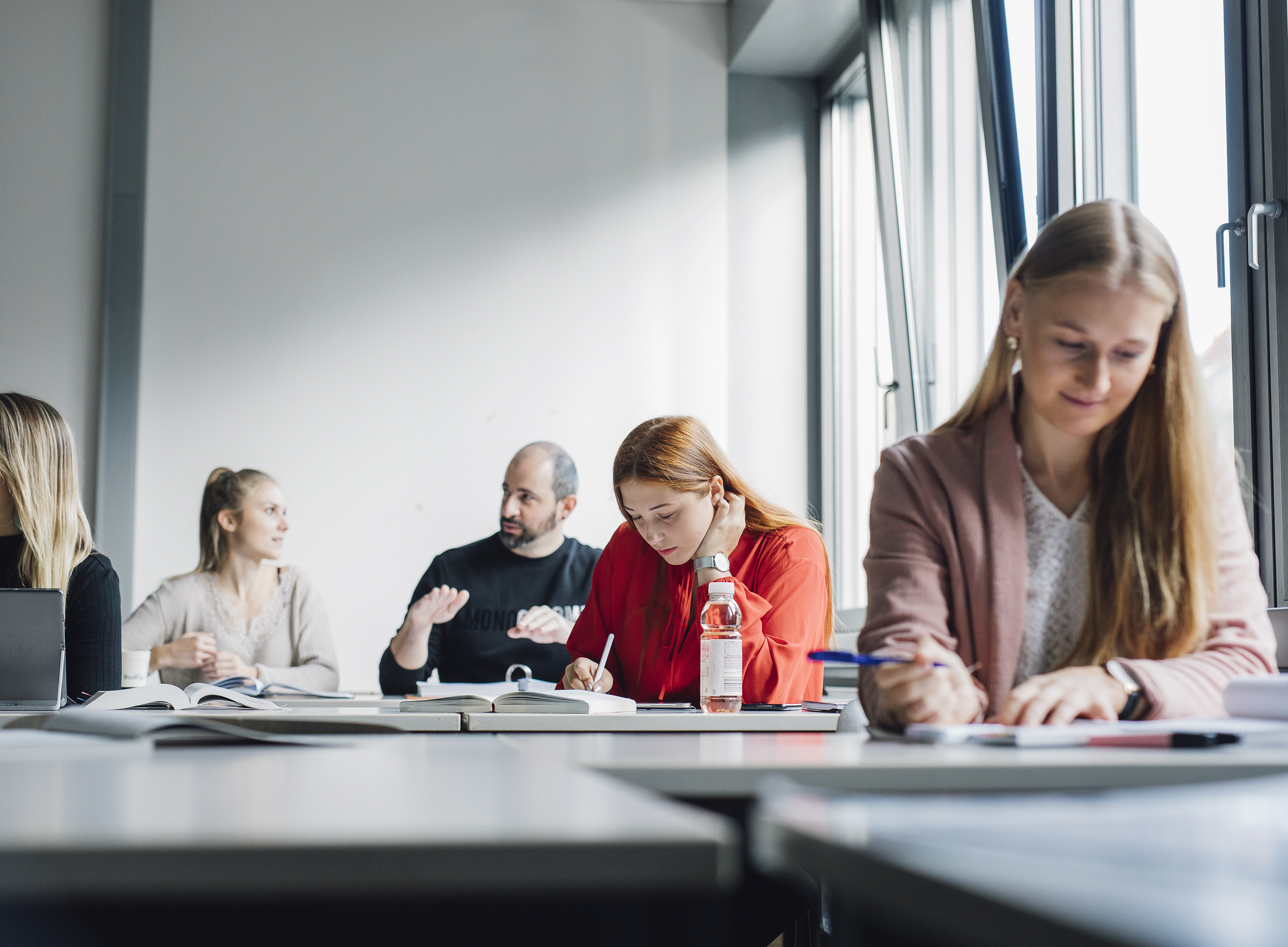 People are sitting at tables near large windows in a bright seminar room, writing in notebooks and working with documents and laptops; writing materials are in the foreground, a water bottle is in the centre, and books and notes are scattered in the background. 
