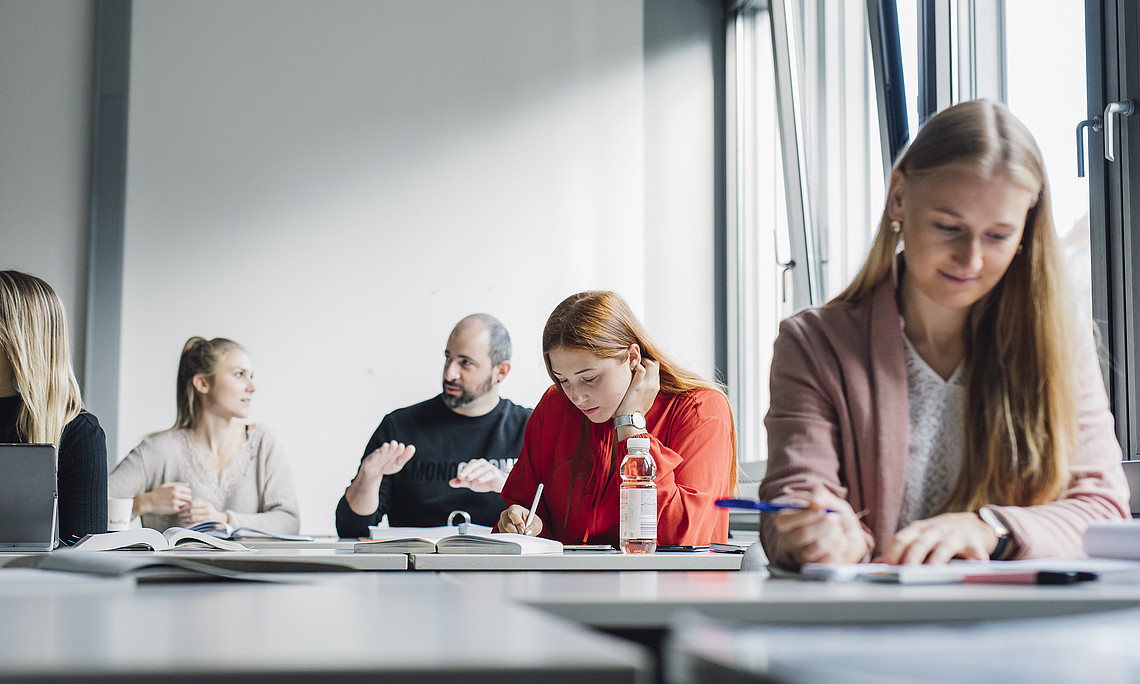 People are sitting at tables near large windows in a bright seminar room, writing in notebooks and working with documents and laptops; writing materials are in the foreground, a water bottle is in the centre, and books and notes are scattered in the background.