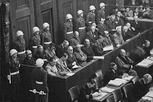 Black and white photograph of a courtroom during the Nuremberg trials. Defendants sit in the upper row, guarded by soldiers with helmets. In front of them are defence lawyers and other people at tables with documents.