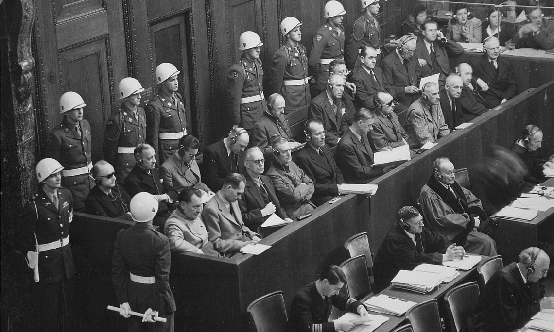 Black and white photograph of a courtroom during the Nuremberg trials. Defendants sit in the upper row, guarded by soldiers with helmets. In front of them are defence lawyers and other people at tables with documents.