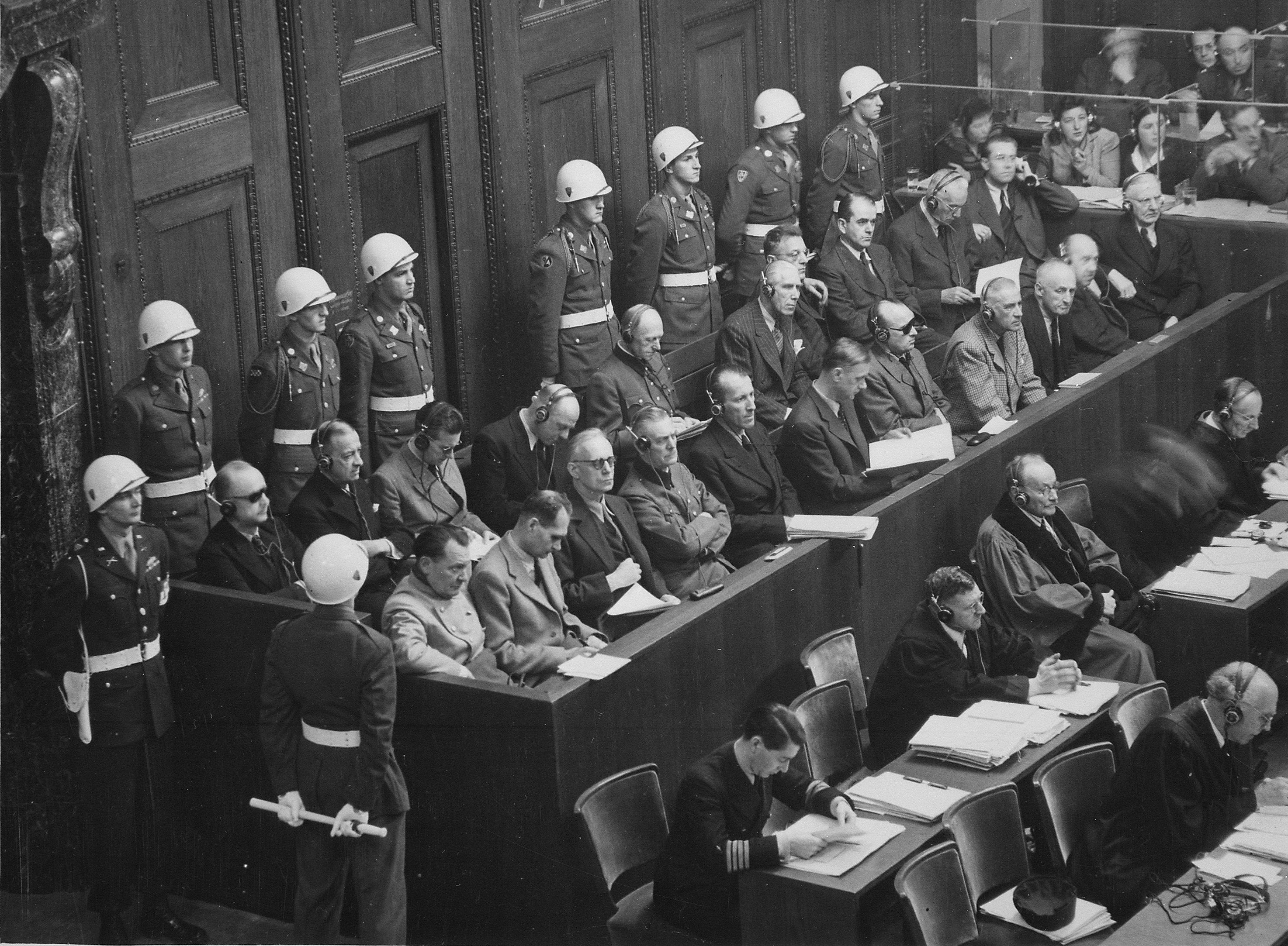 Black and white photograph of a courtroom during the Nuremberg trials. Defendants sit in the upper row, guarded by soldiers with helmets. In front of them are defence lawyers and other people at tables with documents. 