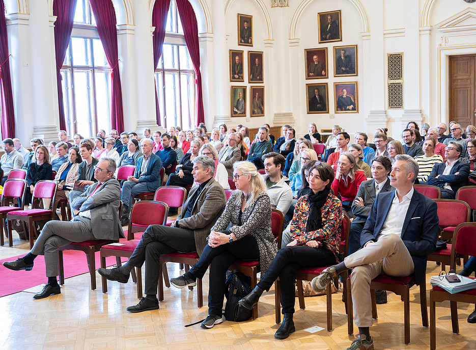In einem großen, historischen Saal sitzen zahlreiche Personen auf roten Stühlen und hören aufmerksam einer Veranstaltung zu. Der Raum wirkt hell und repräsentativ, mit hohen Fenstern und bordeauxroten Vorhängen, weißen Wänden und Stuckverzierungen. An der rechten Wand hängen mehrere gerahmte Porträts. Die Teilnehmenden sind in Alltags‑ bis Businesskleidung gekleidet und schauen überwiegend nach vorne in Richtung einer nicht sichtbaren Bühne oder Redner*in. Auf dem Parkettboden sind zwischen den Stuhlreihen einzelne Taschen und Unterlagen zu sehen. ©Uni Graz/Angele