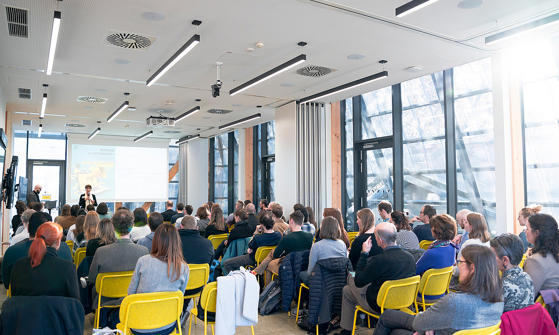 A seated room filled with people looking at a lady on the podium.