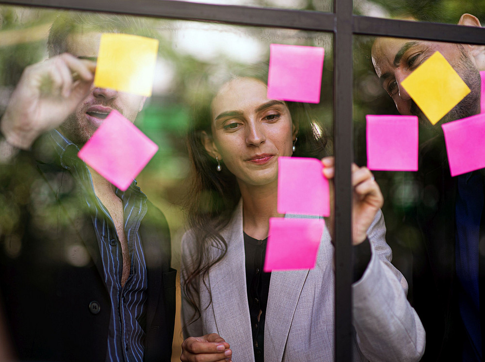 Three people write something down on pink and yellow notes on a glass wall, symbolizing the teaching in the Department of General Education ©SOMPONG DANKHETDAN - stock.adobe.com