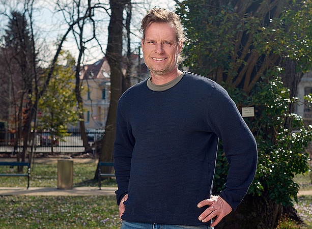 Mann mittleren Alters mit dunkelblauem Pullover und Jeans steht im Park vor Bäumen, Hände in den Hosentaschen, sonniges Wetter. ©Uni Graz / wildundwunderbar