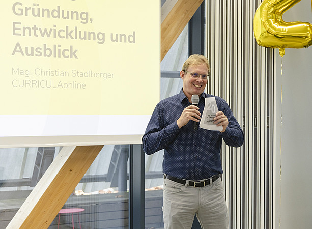 Ein lächelnder Mann hält bei einer Veranstaltung ein Mikrofon und spricht zum Publikum. Auf der Präsentationsfolie steht „Gründung, Entwicklung und Ausblick“. ©@foto-gasser.at