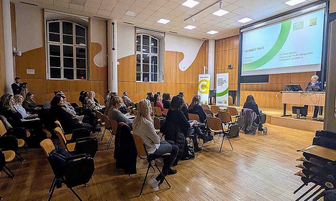 Ein Saal mit hohen Decken, rechts ist eine kleine Bühne mit einer offenen Präsentation auf einer Leinwand darüber und links sind einige Stuhlreihen, und viele Personen, vor allem Frauen, die auf diesen sitzen