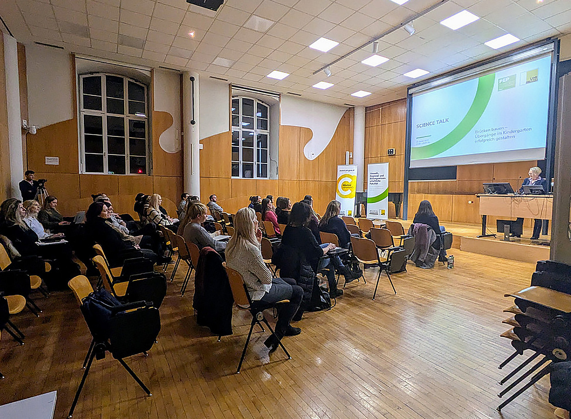 Ein Saal mit hohen Decken, rechts ist eine kleine Bühne mit einer offenen Präsentation auf einer Leinwand darüber und links sind einige Stuhlreihen, und viele Personen, vor allem Frauen, die auf diesen sitzen ©Mateja Marsel