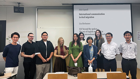 A group of students and professors are standing in a classroom. Julia Gspandl is standing in the middle. Behind them is a projection screen. The screen reads: "Julia Gspandl: International communication in deaf migration. Lisa Bierbaumer: ELF beyond the spoken".