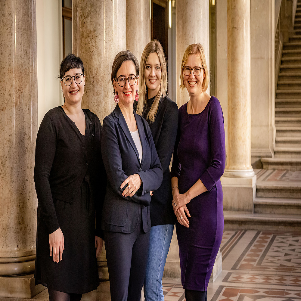 The picture shows four people smiling and posing, probably for a group photo. They are standing in a building with classical architecture, recognizable by the columns and the mosaic floor. From left to right, you can see one person with short dark hair and a black outfit, the second person is wearing glasses and a dark blue blazer over black pants, the third person has medium-length hair and is wearing a gray jacket and jeans, and the fourth person is wearing a purple blouse and black pants. They appear to be in a formal or professional setting and look very content and connected. ©Christine Hofer-Lukic; 15. Österreichischer Zeitgeschichtetag