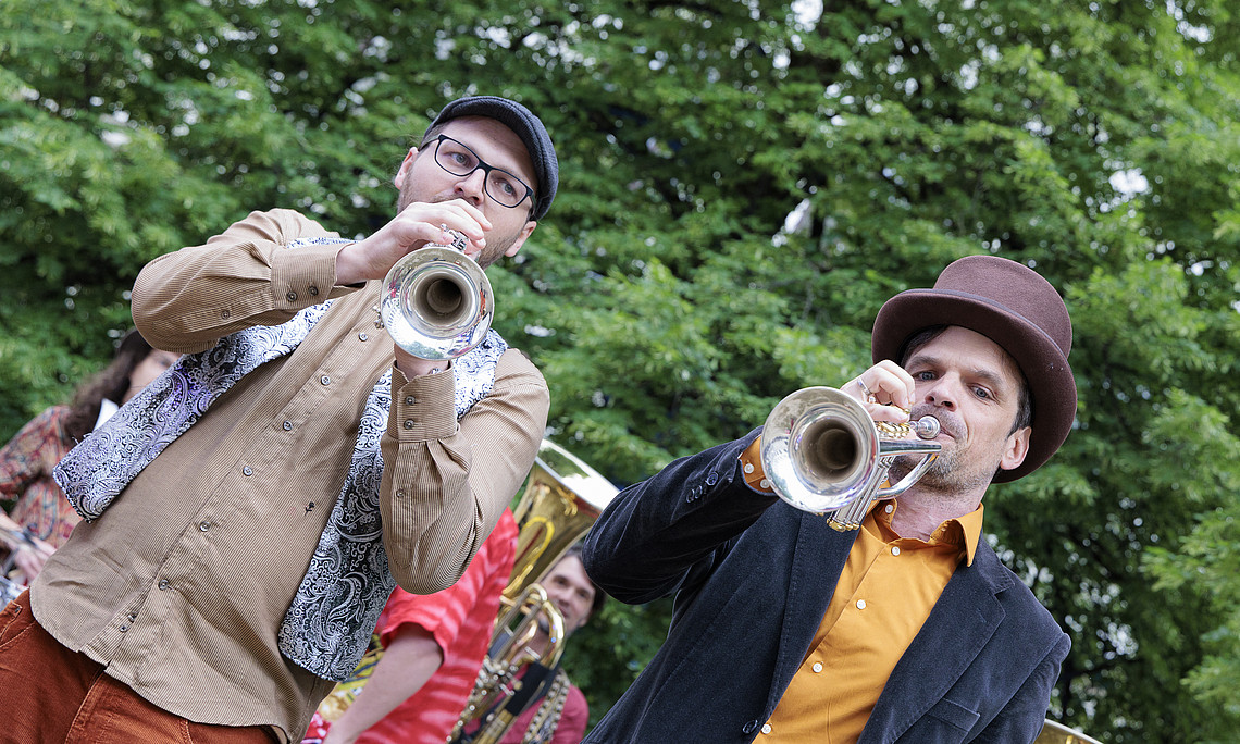 Zwei Männer im Park, einer spielt Posaune, der andere Tuba, beide tragen braune Fedora-Hüte, sind leger gekleidet und stehen auf einer Party im Freien vor dem Hintergrund grüner Bäume.