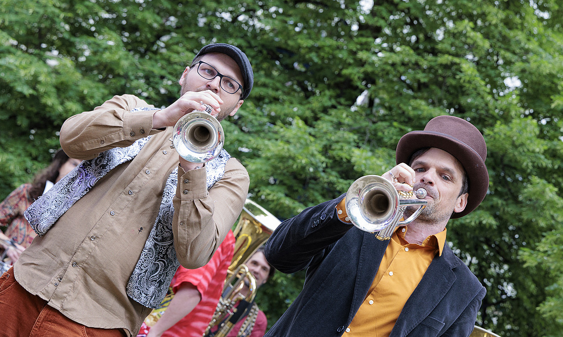 Two men in the park, one plays a trombone and the other a tuba, both wearing brown fedora hats, casual , at an outdoor party, with a background of green trees.
