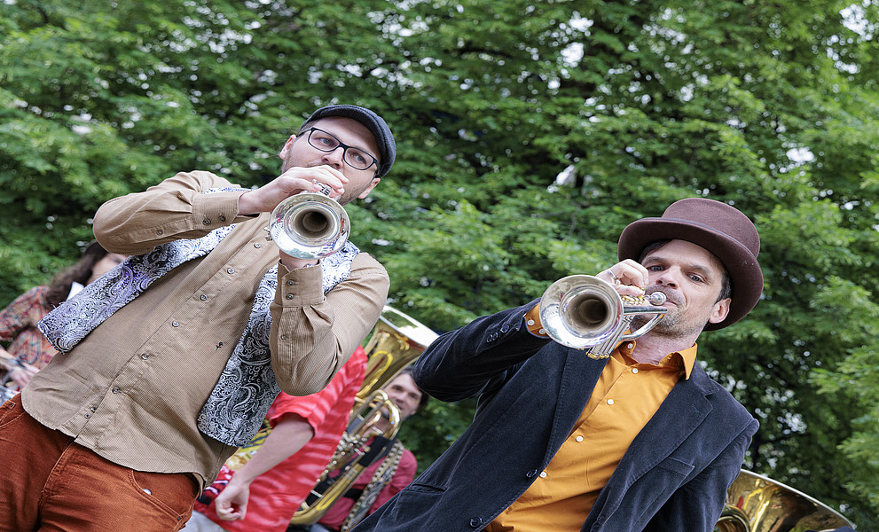 Zwei Männer im Park, einer spielt Posaune, der andere Tuba, beide tragen braune Fedora-Hüte, sind leger gekleidet und stehen auf einer Party im Freien vor dem Hintergrund grüner Bäume.