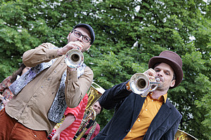 Two men in the park, one plays a trombone and the other a tuba, both wearing brown fedora hats, casual , at an outdoor party, with a background of green trees.