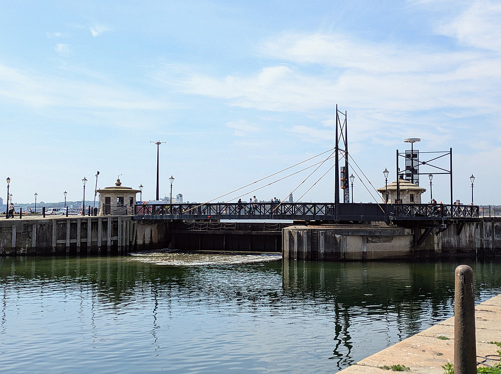 Blick auf eine historische Drehbrücke, die ein ruhiges Hafenbecken in Liverpool überspannt. Die Brücke hat schwarze Geländer und Seile und verbindet zwei kleine Kontrolltürme aus Stein mit gewölbten Dächern. Unter strahlend blauem Himmel gehen mehrere Menschen über die Brücke, und das Wasser darunter spiegelt die Konstruktion und die umliegenden Lampen entlang des Kais wider. ©Daniella Shaw