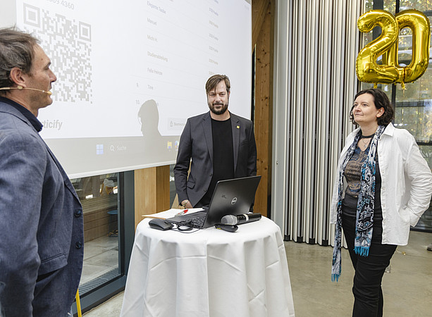 Zwei Männer und eine Frau stehen bei einem Stehtisch. Im Hintergrund zeigt eine Präsentation einen Bildschirm mit Quizfragen; goldene Luftballons bilden die Zahl 20. ©@foto-gasser.at