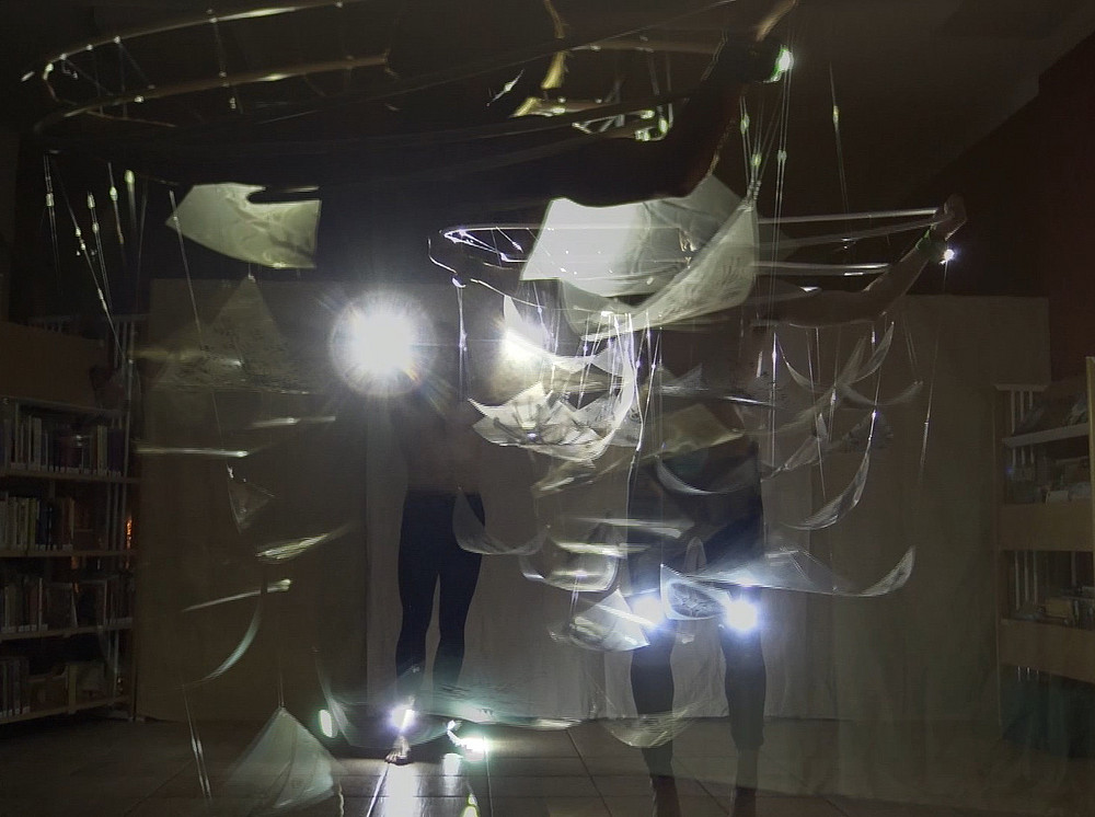 Das Foto zeigt zwei Personen in einer schwach beleuchteten Bibliothek, die Lichter halten, die transparente, wirbelnde Blätter beleuchten, die um sie herum aufgehängt sind. Im Hintergrund sind Bücherregale zu sehen. ©Oksana Chepelyk