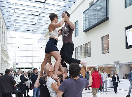 Foto von zwei Männern und einer Frau, die in einem Flur zwischen zwei Gebäuden eine akrobatische Tanznummer aufführen, während eine Menschenmenge ihnen zusieht. ©Uni Graz/Schweiger