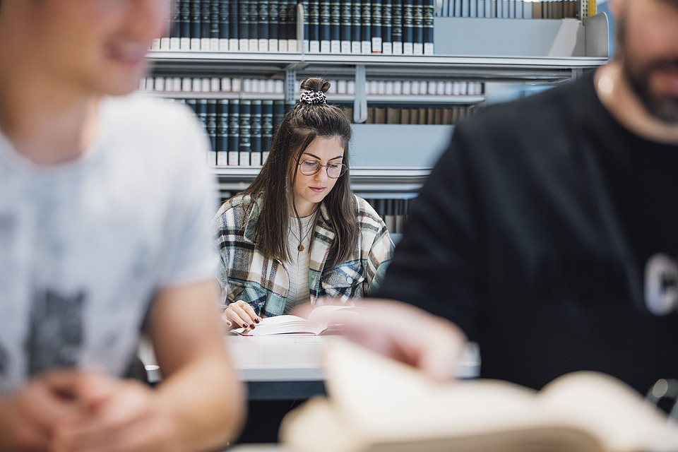 Eine Studierende sitzt in der Universitätsbibliothek mit einem Buch an einem Tisch vor einem Bücherregal; vor ihr sind verschwommen zwei weitere Studierende zu sehen.