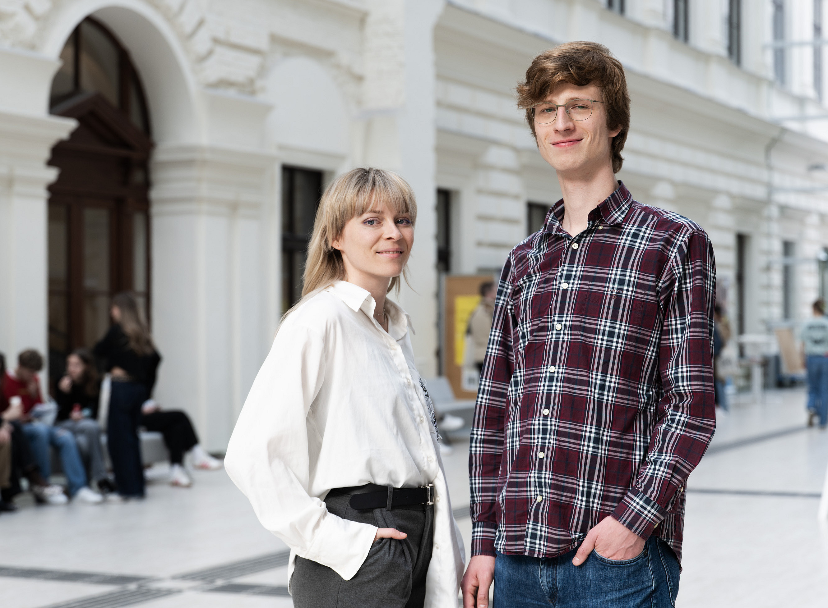 Zwei Personen stehen in einem hellen, offenen Innenraum mit hohen Decken und großen Fenstern. Die Person links trägt ein weißes Hemd und eine graue Hose, während die Person rechts ein rot-kariertes Hemd und eine blaue Jeans trägt. Im Hintergrund sind weitere Personen auf einer Sitzbank sowie architektonische Details wie Bögen und Fenster zu sehen. 