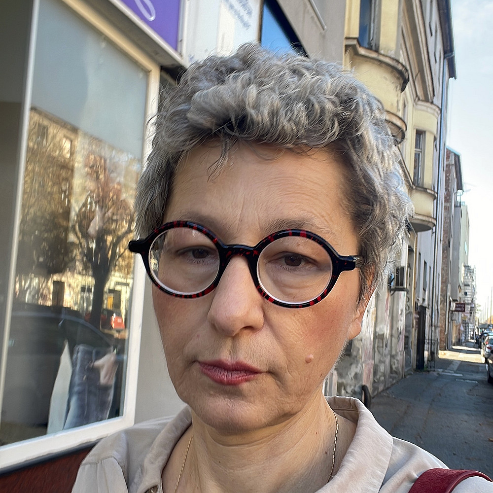 Porträt von Milka Car, einer Frau mit kurzen grauen Haaren und runder Brille, die frontal in die Kamera blickt. Sie trägt eine helle Bluse und eine Schultertasche und steht auf einem Gehweg vor Gebäuden in einer städtischen Umgebung bei Tageslicht. ©Milka Car