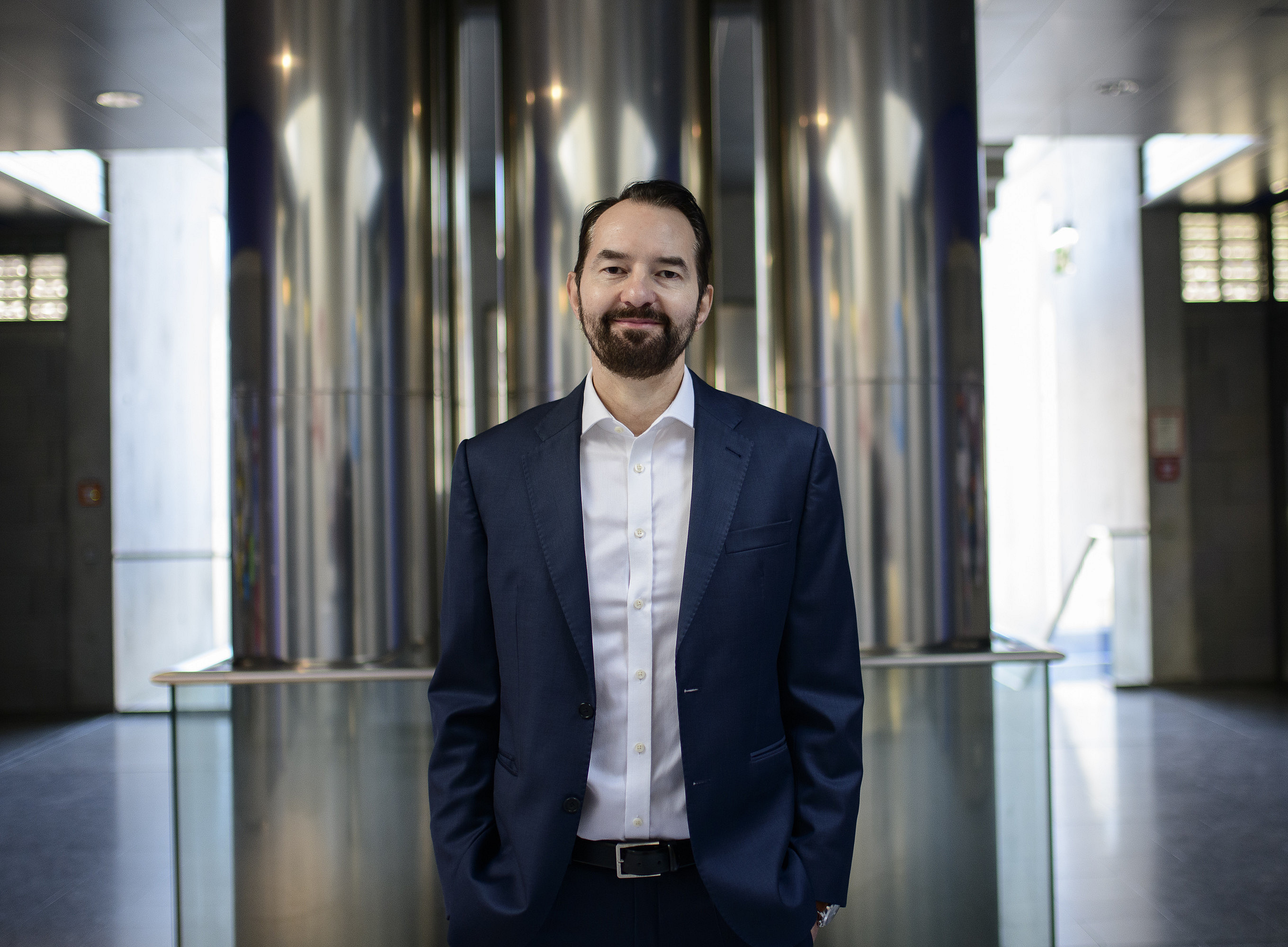 Ein Porträt eines Mannes in Geschäftskleidung, der selbstbewusst in einem Bürogebäude mit Edelstahlsäulen und Glaswänden steht und professionelle Eleganz ausstrahlt. Er hat dunkles Haar und einen Bart, trägt einen blauen Anzug, ein weißes Hemd und schwarze Schuhe. Er lächelt in die Kamera. ©Uni Graz/Tzivanopoulos