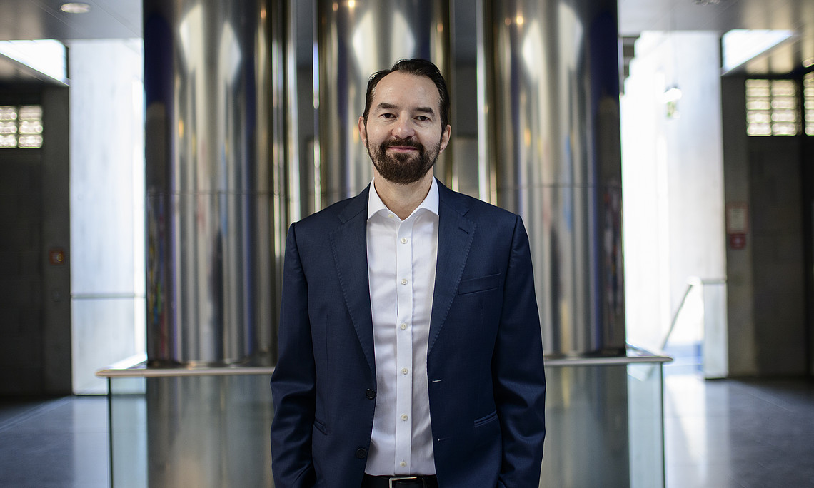 a portrait of a man in business attire, standing confidently inside an office building with stainless steel columns and glass walls, exuding professional elegance. he has dark hair and a beard, wearing a blue suit, white shirt, and black shoes. he is smiling at the camera