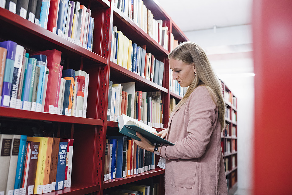 Eine Studentin steht in einer Bibliothek vor einer Bücherwand und liest in einem Buch