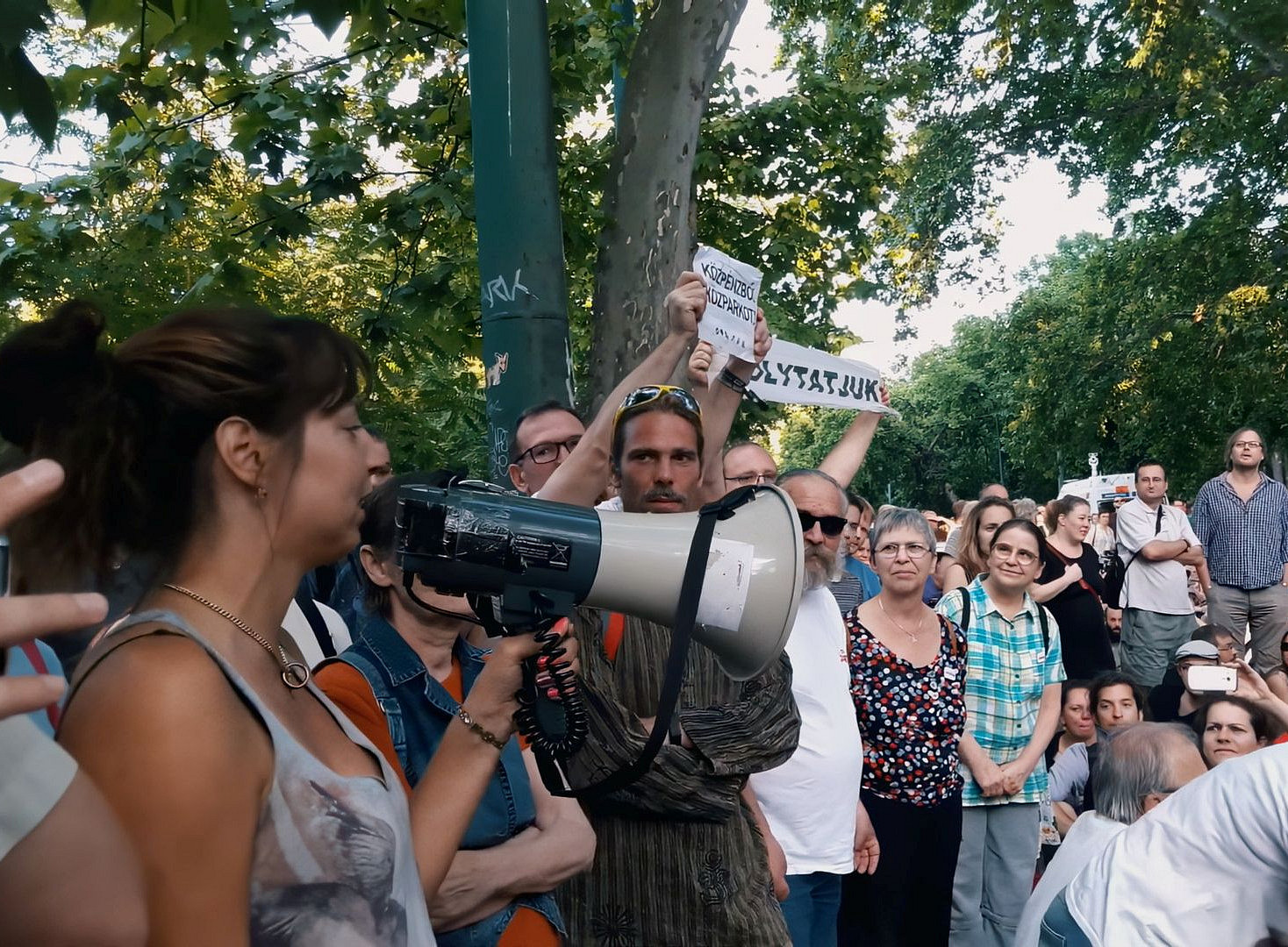 eine Gruppe Menschen bei einer Demonstration im Park: im Vordergrund spricht eine Frau in ein Megaphon, ein paar Menschen fotografieren sie; andere Leute halten Schilder in ungarischer Sprache hoch ©Ádám Csillag