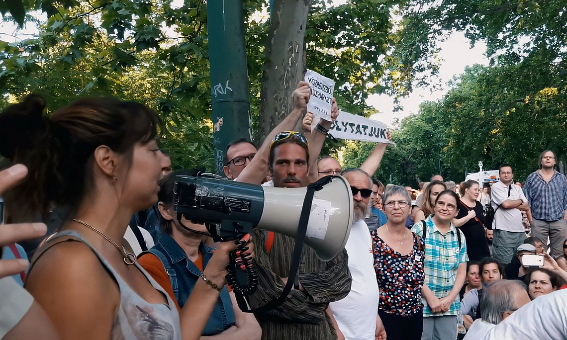 eine Gruppe Menschen bei einer Demonstration im Park: im Vordergrund spricht eine Frau in ein Megaphon, ein paar Menschen fotografieren sie; andere Leute halten Schilder in ungarischer Sprache hoch