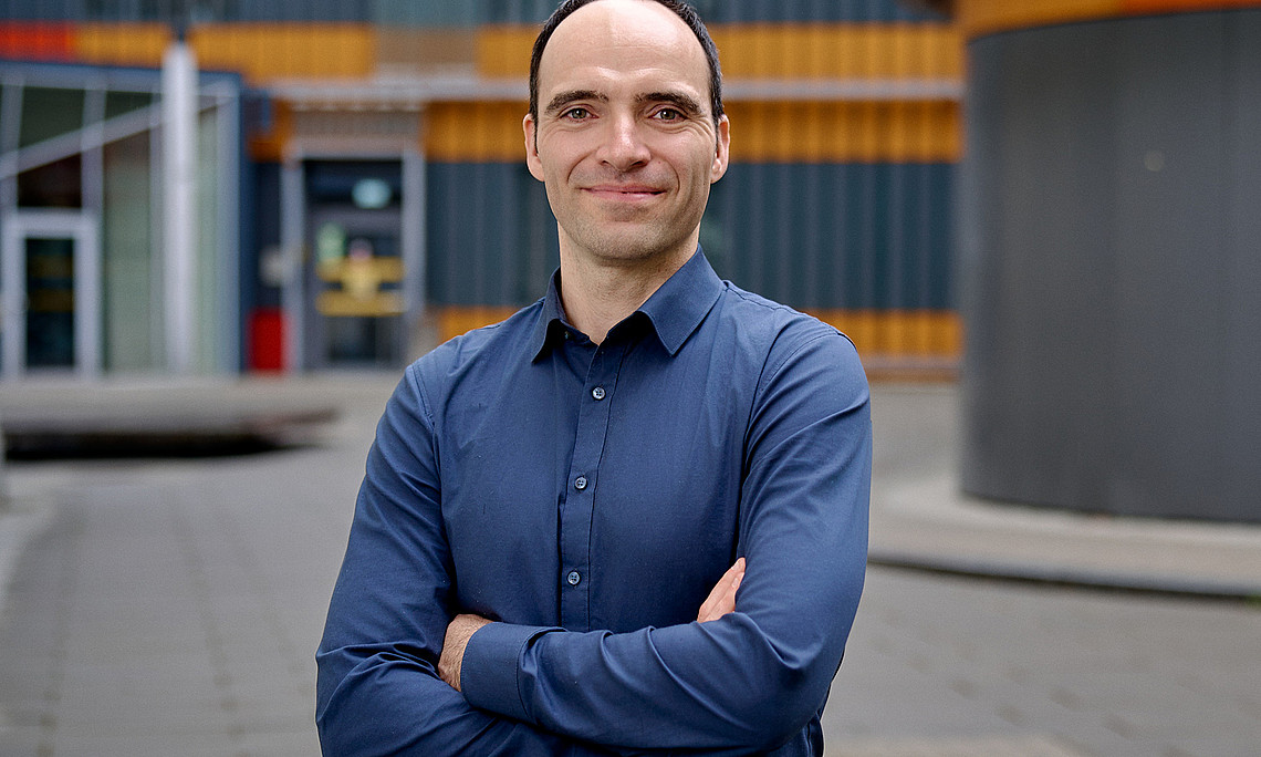 Sebastian Preissl with arms crossed, chest portrait, in the background the Centre for Molecular Biosciences at the University of Graz
