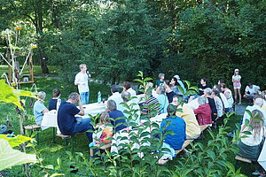 Vortragender steht vor einer Gruppe von Personen, die in einem Garten auf Bänken sitzen und ihm zuhören