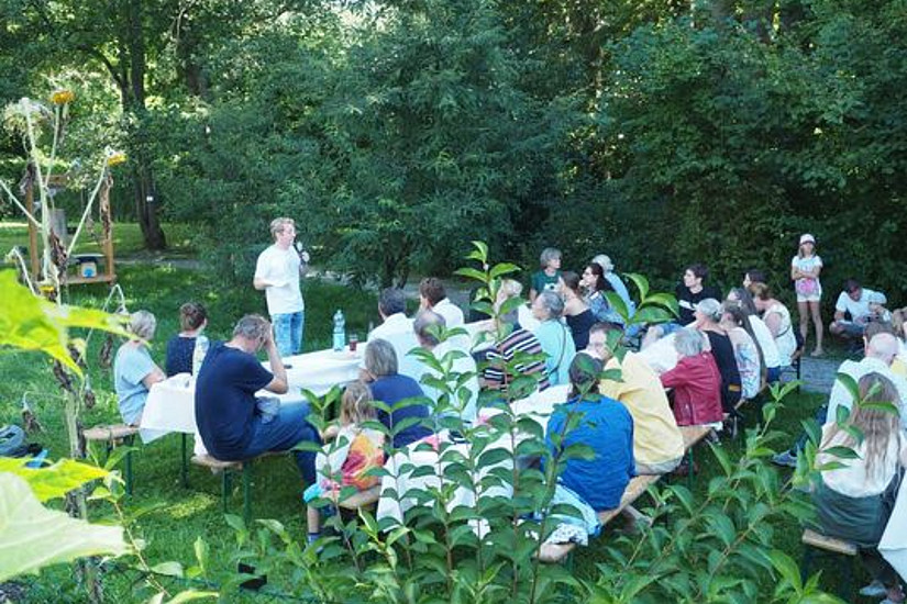 Vortragender steht vor einer Gruppe von Personen, die in einem Garten auf Bänken sitzen und ihm zuhören