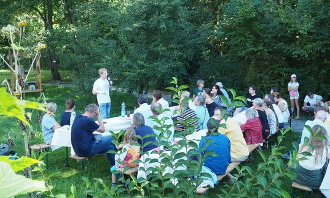 Vortragender steht vor einer Gruppe von Personen, die in einem Garten auf Bänken sitzen und ihm zuhören