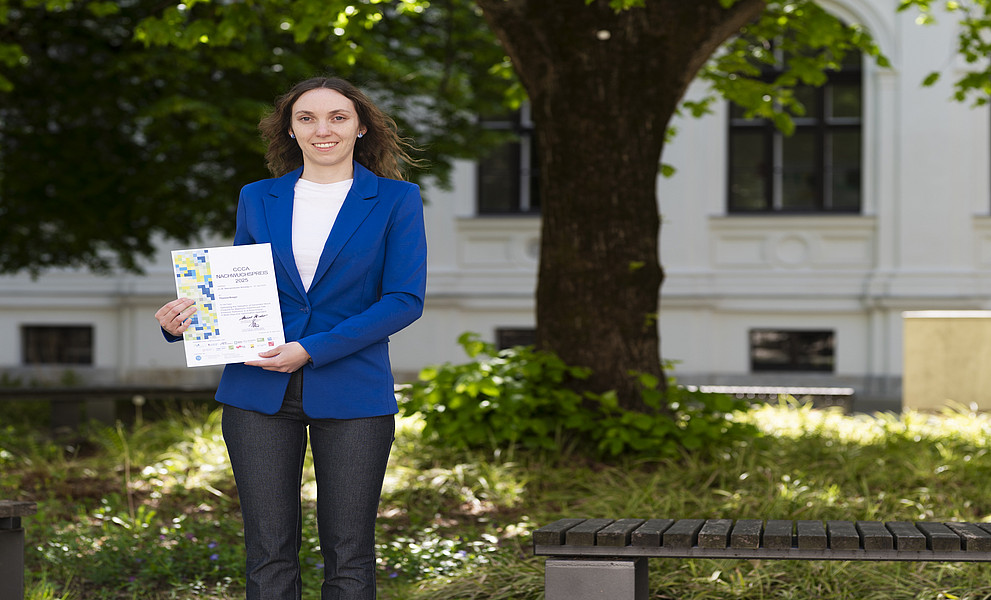 Eine Frau steht im Innenhof des Universitätsgebäude und hält eine Urkunde.