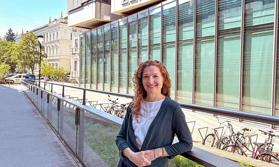 Frau steht vor einem Gebäude mit Glasfassade, lehnt sich an ein Glasgeländer und schaut in die Kamera