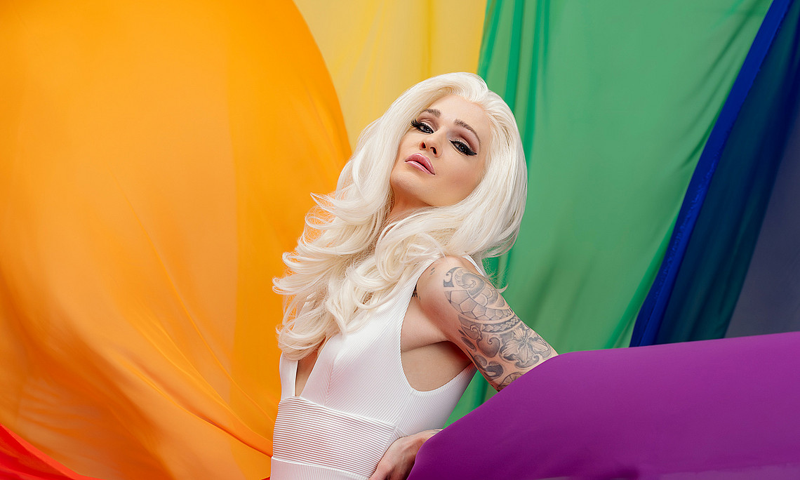 Dragqueen Gloria Hole im weißen Kleid mitten in regenbogenfarbigen Stoffen