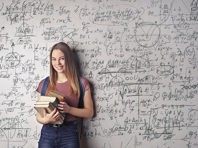 Lächelndes Mädchen mit Schulbüchern in der Hand steht vor einer Tafel mit mathematischen Formeln 