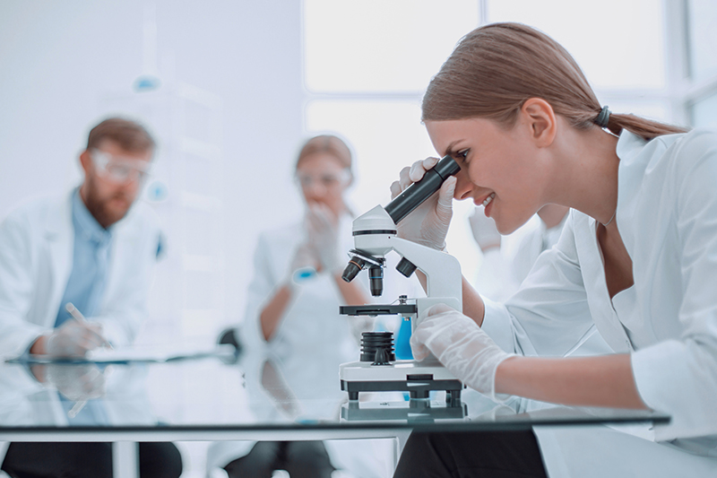 Junge Forscherin schaut in ein Mikroskop, im Hintergrund arbeiten Menschen im Labor 