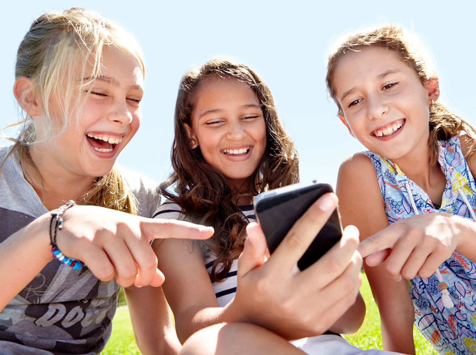 Drei lachende Mädchen, die auf ein Handy schauen. Das Mädchen in der Mitte hält das Handy in der Hand, die beiden anderen zeigen mit einem Finger darauf. ©PeopleImages.com - #52d4e6c251413